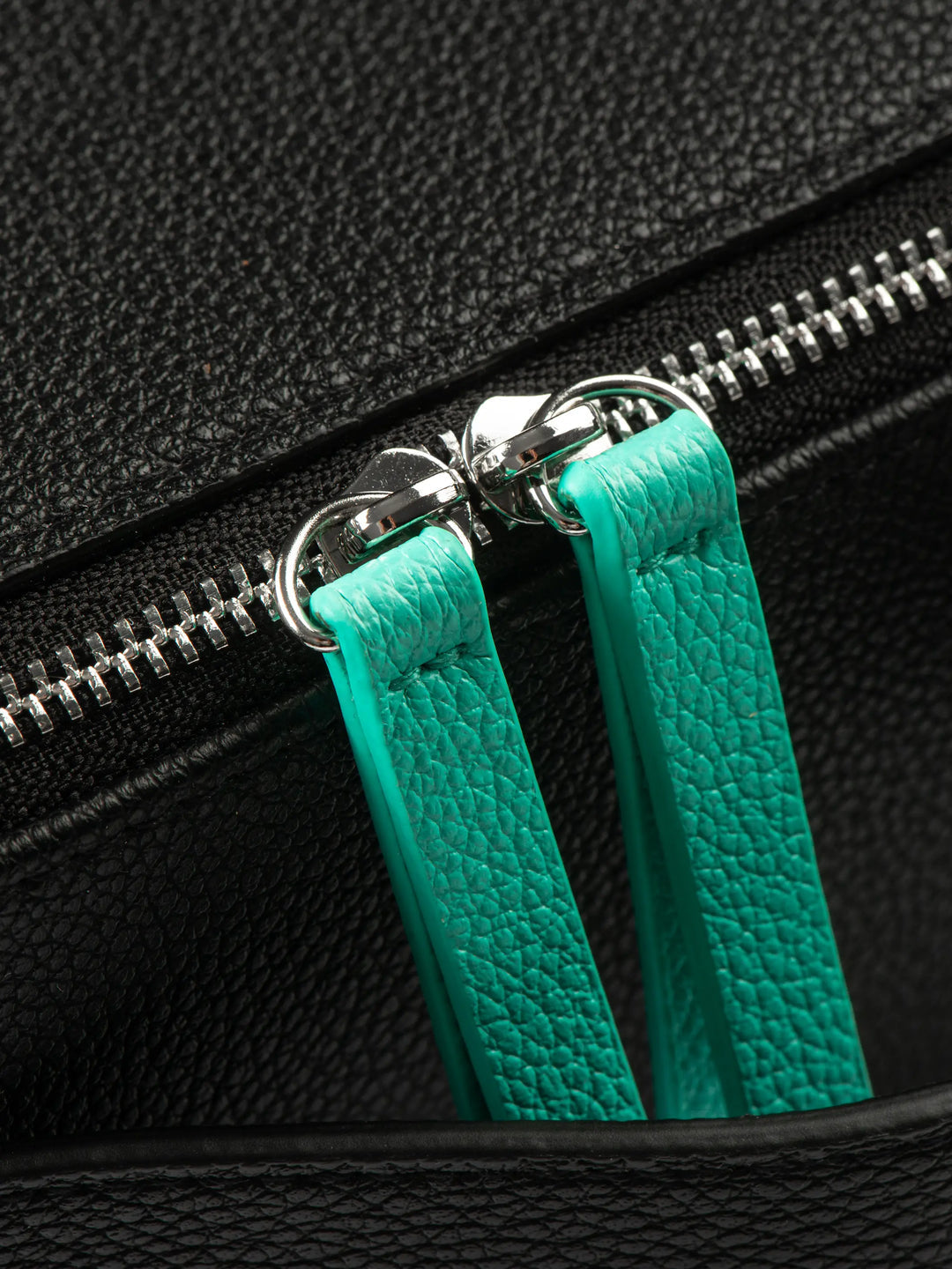 Close-up of a black bag with a silver zipper and green leather pull handles.